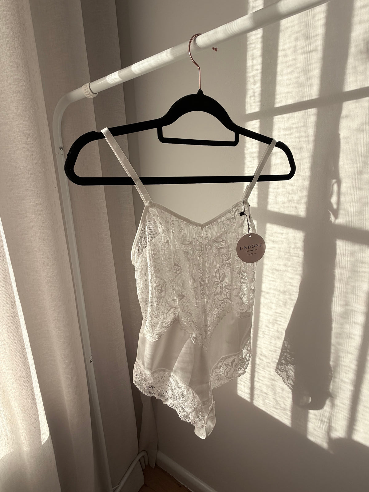 A white lace camisole top hanging on a black hanger from a white clothesline, with strong window shadow patterns cast across the scene.